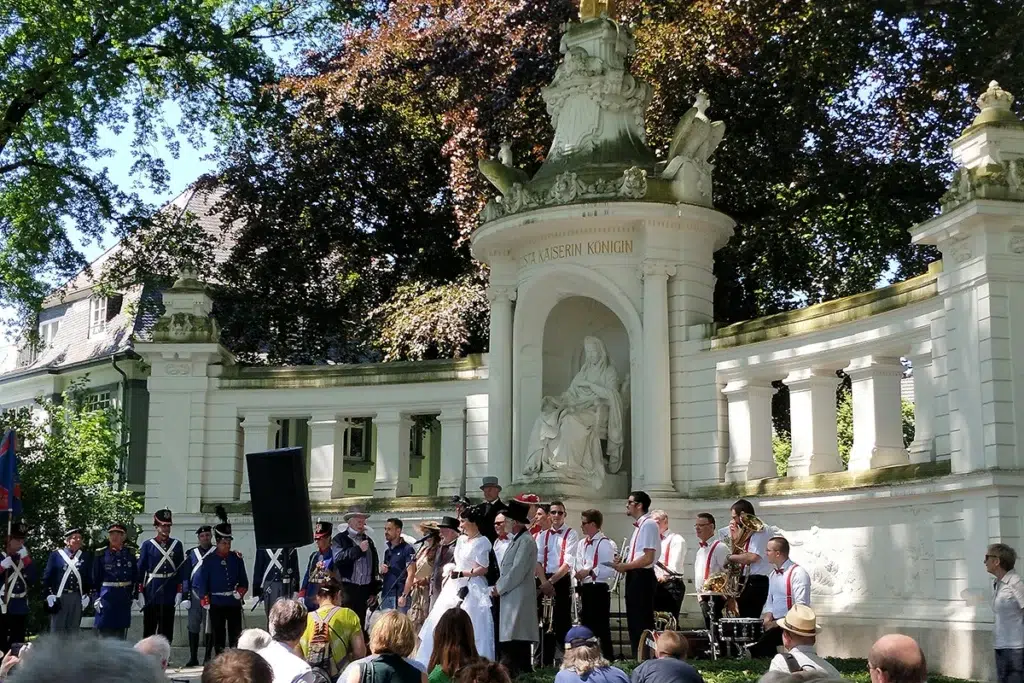 Eine Band und uniformierte Personen treten vor einem Denkmal im Freien mit einer sitzenden Statue auf und werden dabei von einem Publikum beobachtet. Das Denkmal steht vor einer Kulisse aus Bäumen und einem Gebäude im klassischen Stil.