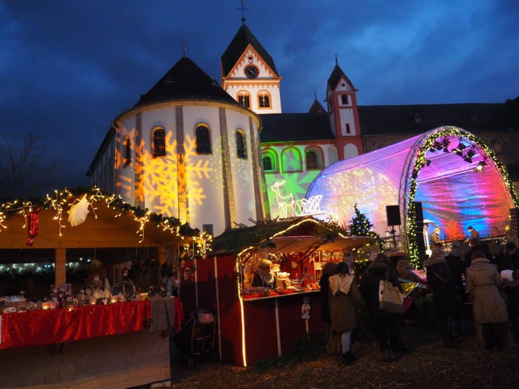 Ein festlicher Markt in der Abenddämmerung mit Ständen, Lichtern und Menschen. Eine Kirche mit bunten Lichtern und Schneeflockenprojektionen im Hintergrund.