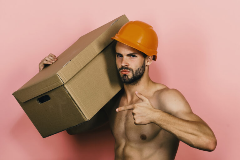 Ein hemdsärmeliger Mann mit orangefarbenem Schutzhelm trägt einen großen Karton auf der Schulter und zeigt auf ihn, während er vor einem rosafarbenen Hintergrund steht.