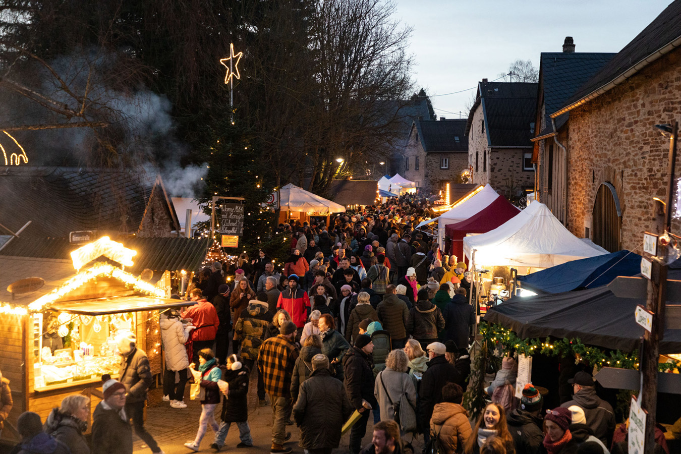 Eine Menschenmenge schlendert zwischen festlichen Ständen und Lichtern auf einem abendlichen Weihnachtsmarkt im Freien.