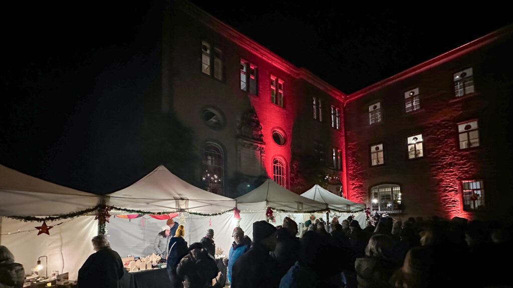 Nächtlicher Markt unter freiem Himmel mit weißen Zelten, Menschen, die sich an den Ständen umsehen, und einem mit roten Lichtern beleuchteten Backsteingebäude im Hintergrund.