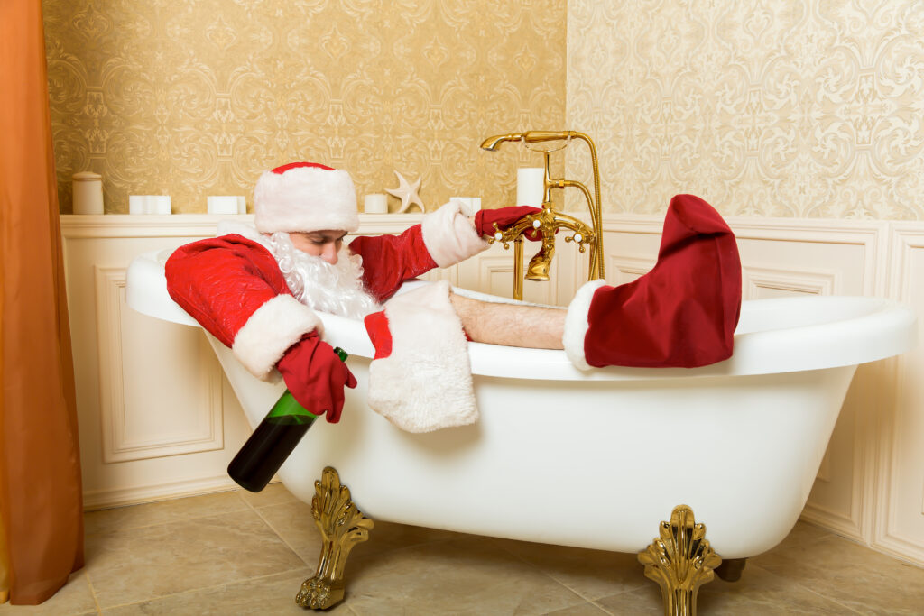 Eine Person im Weihnachtsmannkostüm liegt in einer weißen Badewanne mit Krallenfuß, hält eine Flasche in der Hand und wirkt entspannt, mit goldenen Armaturen und dekorativen Kerzen im Hintergrund.