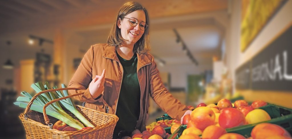 Eine Frau mit Brille und in einer braunen Jacke lächelt, während sie auf einem Hallenmarkt Äpfel aussucht; in der Hand hält sie einen Korb voller Lauch.