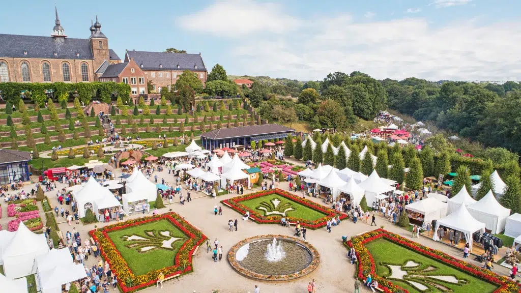Luftaufnahme eines Gartenfestes mit weißen Zelten, einem Springbrunnen in der Mitte, Blumenbeeten und einem historischen Gebäude im Hintergrund. Menschenmengen schlendern zwischen den Ausstellungsständen und dem Grün herum.